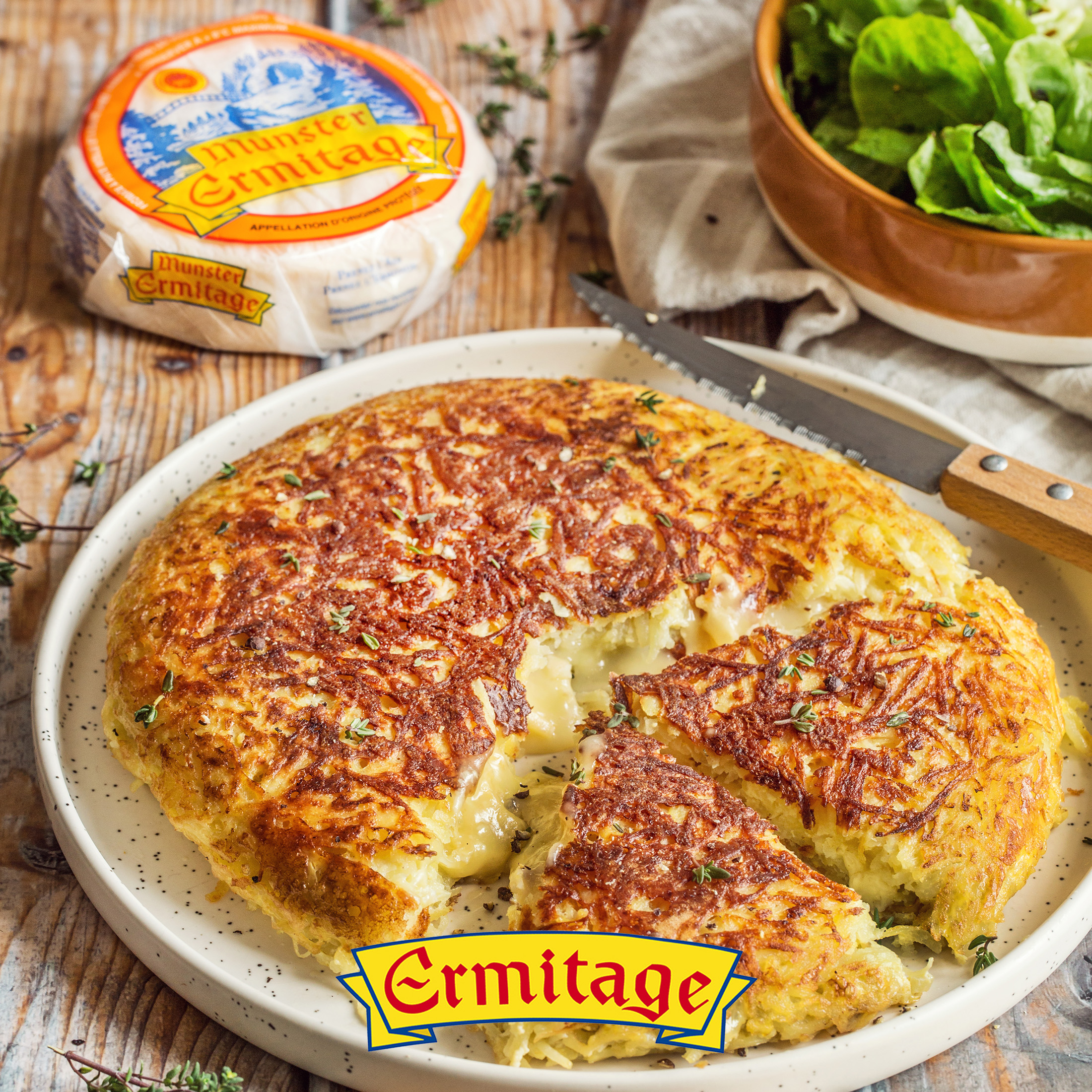 Grand rösti au Munster Ermitage et oignons, coupé en parts dans une assiette, avec une salade verte.